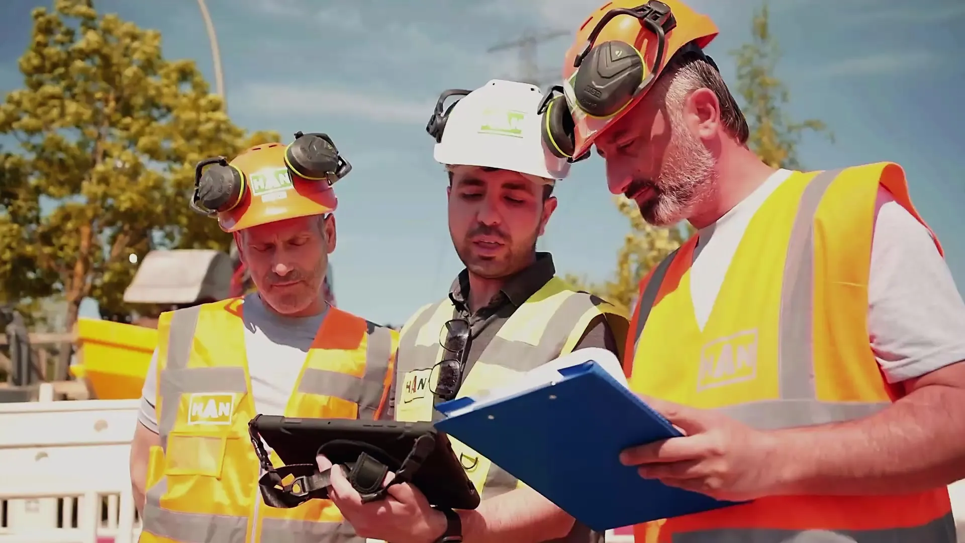 Drei HAN-Mitarbeiter mit Helm und Warnweste stimmen auf der Baustelle den Ablauf mit Tablet und Klemmbrett ab